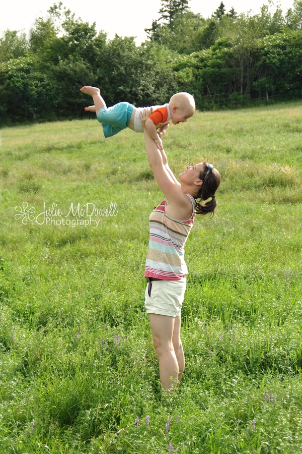 mother in striped tank top in field lifting young toddler over her head